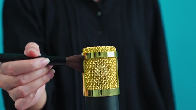 Close-up of Hand brushing gold condenser microphone with makeup brush on cyan background
