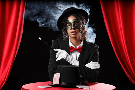 Young African-American magician in mask with magic wand, hat and cards sitting at table on black background