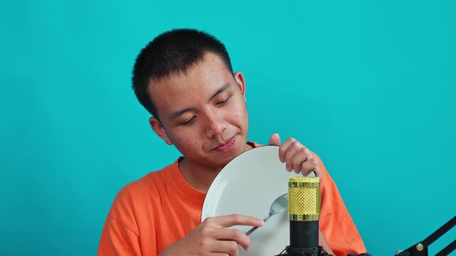 Asian man scraping fork on ceramic plate near gold condenser microphone on cyan background