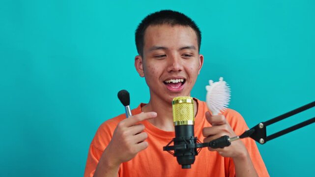Asian man waving and holding props near gold condenser microphone on cyan background