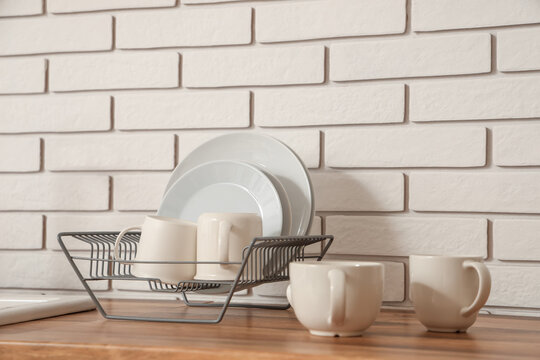 Rack with clean plates and cups on wooden table near light brick wall