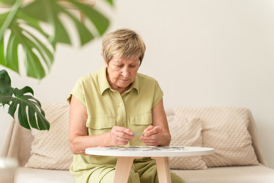 Elderly woman sits on sofa at table, intently working on puzzle, engaging in leisure activities for mental stimulation and brain health. A bright, cozy home interior