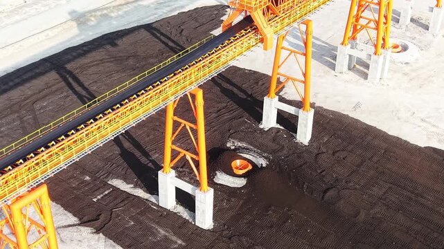 High-angle aerial 4K video of a bright orange industrial conveyor belt system operating across a large dark coal stockpile and concrete infrastructure yard.