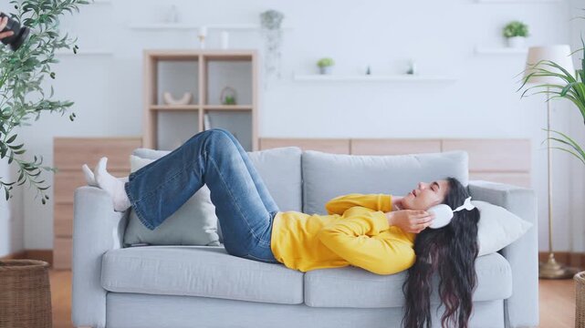 Asian woman is listening to music or a podcast through headphones to relax in the living room. Young lady is enjoying lounging on the sofa at home. Meditation for mental health and wellbeing
