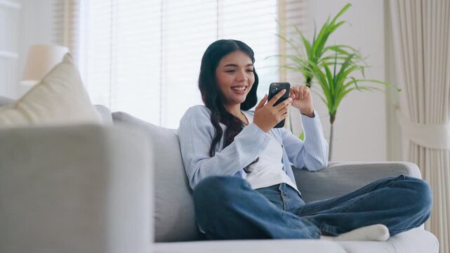 Happy young asian woman relax on comfortable couch at home texting messaging on smartphone, smiling girl use cellphone, chatting online message, shopping online from home