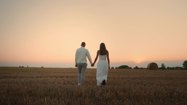 Romantic couple walking hand in hand across a tranquil harvested field at sunset, silhouettes framed by golden sky and distant hay bales in peaceful countryside landscape