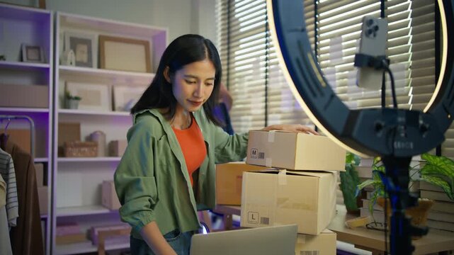 Young Asian woman fashion entrepreneur live streaming selling clothes online with smartphone ring light and laptop in home studio, representing small business owner and professional women.