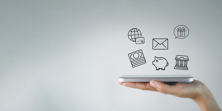 Hand holding a smartphone with financial and banking overlay icons on a grey background. Concept of mobile banking, fintech, online shopping, digital wallet, and saving money with copy space.