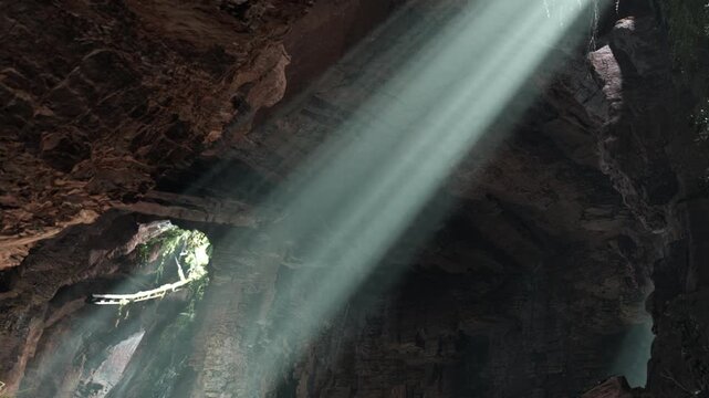 Sunlight shines through cracks in the cave ceiling. The rocks around are dark and rough. The light creates a natural spotlight effect inside the cave.