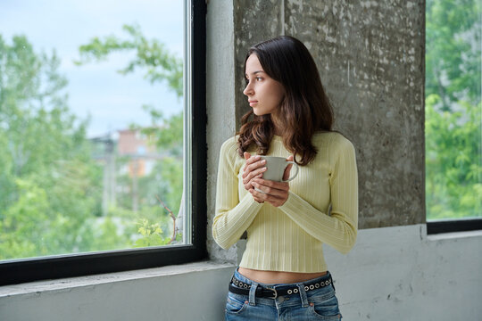 Pensive young woman holding mug standing near window and looking outside