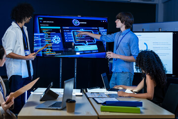 A junior programmer wearing round glasses walks his colleagues through the new interface in a dark...