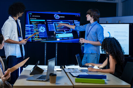 A junior programmer wearing round glasses walks his colleagues through the new interface in a dark blue lit technology office at night, an image of a young talent finding his voice