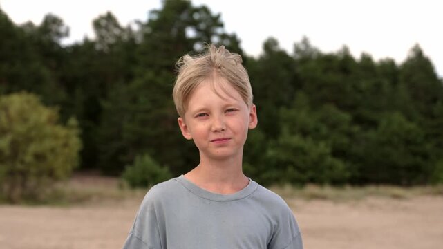 Portrait of a cheerful young caucasian boy with blond hair laughing and smiling genuinely at the camera, with a calm expression at the end, standing in a natural park with a sandy beach and forest