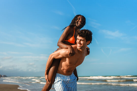 Young happy interracial couple enjoying a piggyback ride by the ocean on a summer day