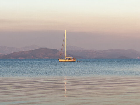Anchored sailboat on calm Garitsa Bay at pastel sunset, Corfu Town, Greece, hazy horizon