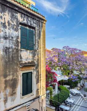Weathered facade with green shutters, purple blooms, cafe tables in Corfu Old Town, Greece