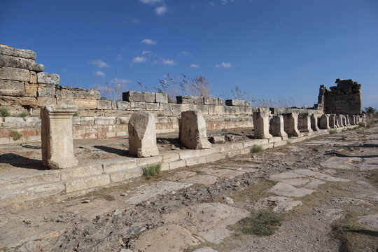 "Remains of an ancient structure in Ephesus, featuring a weathered stone wall, fallen column bases, and dry grasses under a blue sky