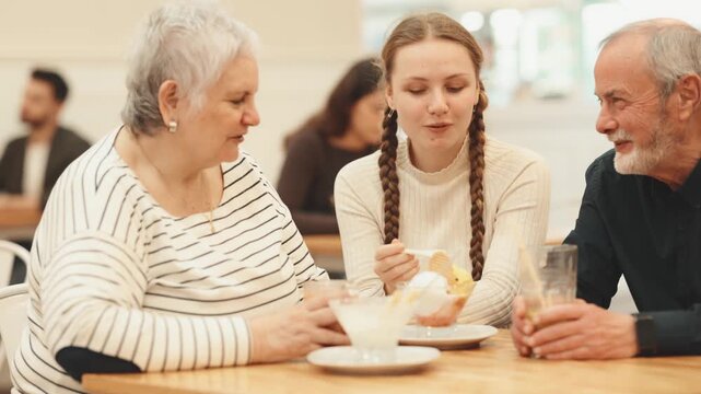 Teenage girl is talking with interest to her grandparents in a cafe, they ordered drinks with desserts. High quality 4k footage