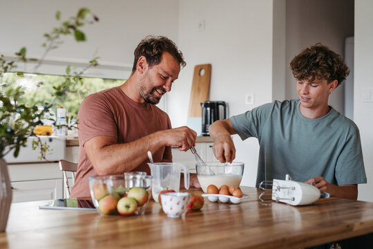 Dad teaching teen son how to bake cake at home.