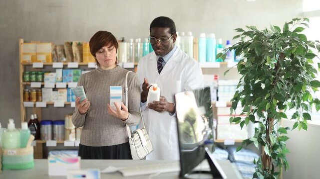 Woman choose buy painkiller tablet at pharmacy, compare with other good. Client check for absence of harmful substances, view product in details. African man pharmacist offer available assortment item