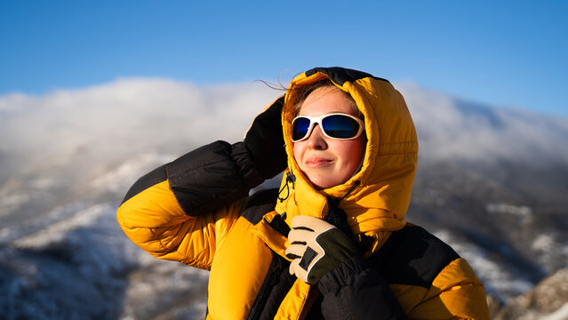 Portrait of a woman in a warm yellow jacket and protective sunglasses standing in sunlight in high mountains under a clear blue sky.