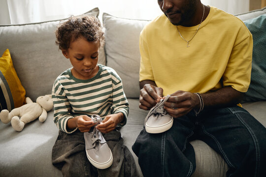 African American father helping male child tying shoelaces indoors. Concept depicts early childhood development, ideal for parenting education, family lifestyle marketing