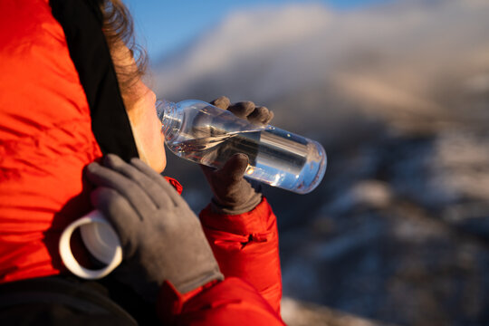 Female mountaineer drinking water from a reusable bottle during a winter mountain ascent at sunrise, surrounded by snowy peaks.