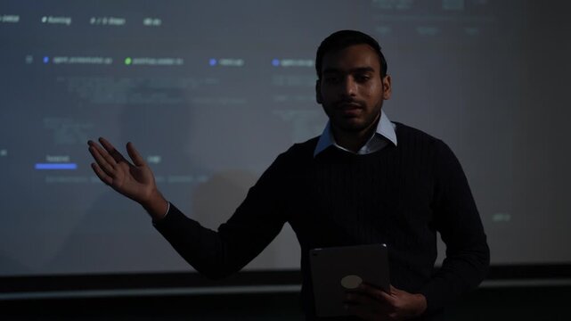 A bearded South Asian AI engineer in a navy knit sweater holds a tablet and opens one palm as he explains an LLM agent orchestration dashboard projected behind him in a dark tech office.