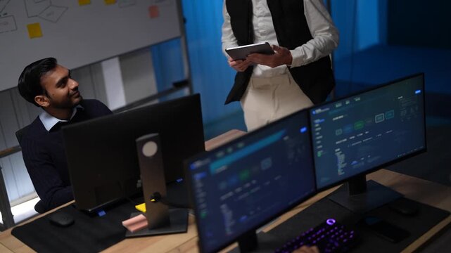 A team lead in a black vest holding a tablet leans over a workstation to brief a seated engineer about the metrics on the monitors in front of them in a dark tech office.