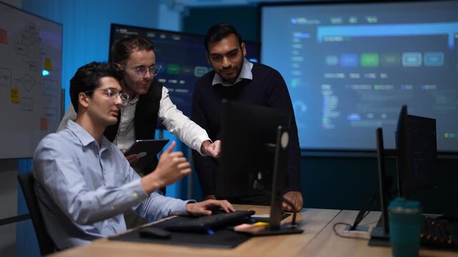 A programmer at his keyboard with two colleagues behind him gets a laugh from the group as the run they were waiting for finally turns green on his screen in a dark tech office.