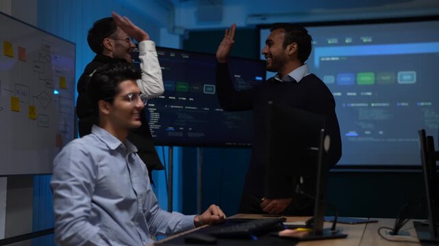 Two standing colleagues over a seated developer throw a celebratory high five above the desk the moment their model deployment succeeds inside a dark AI startup office.