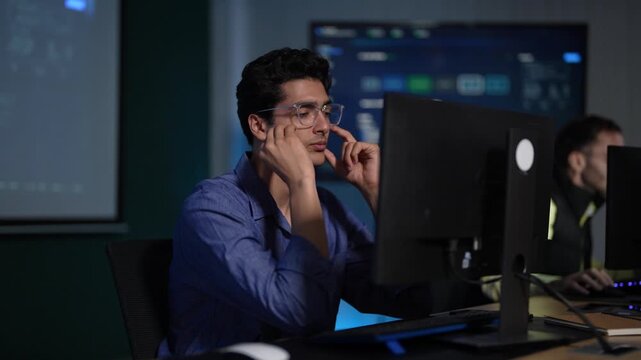 A young engineer in a blue shirt and glasses rests his fingers against his temple and stares at a stubborn problem on the monitor in front of him in a dark tech office.