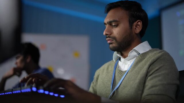 A developer in an olive sweater and lanyard keeps typing while a colleague's hand gestures across his screen and a third teammate works behind in a dark tech office.