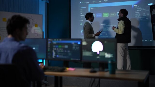 An engineer reaching for his keyboard keeps working as the pair behind him trade points in front of the projected system map inside a dimly lit machine learning startup office.