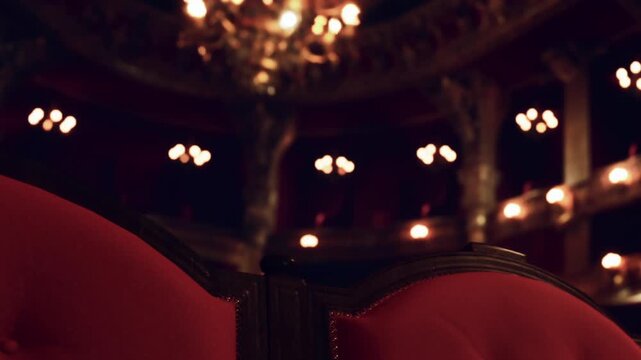 Red upholstered chairs are visible in a theater setting. The background features dim lighting and decorative architecture. The atmosphere feels lively and full of anticipation during a performance.