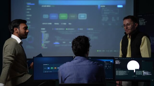 A technical founder addressing his team gestures with an open hand mid sentence while a listener sits silhouetted in the foreground inside a dark AI startup office.