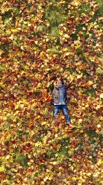 Vertical video: Aerial view of a happy little girl lying on yellow autumn leaves in park, dreamily looking up at camera. Social media format.