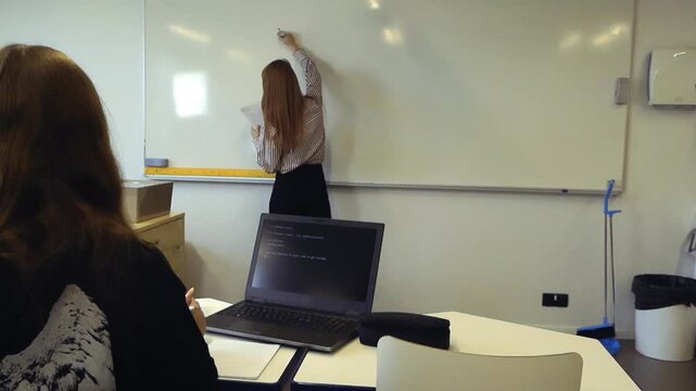 Female teacher writing a math problem on the whiteboard for a young student sitting at her desk with a laptop, which is showing lines of code for an artificial intelligence program