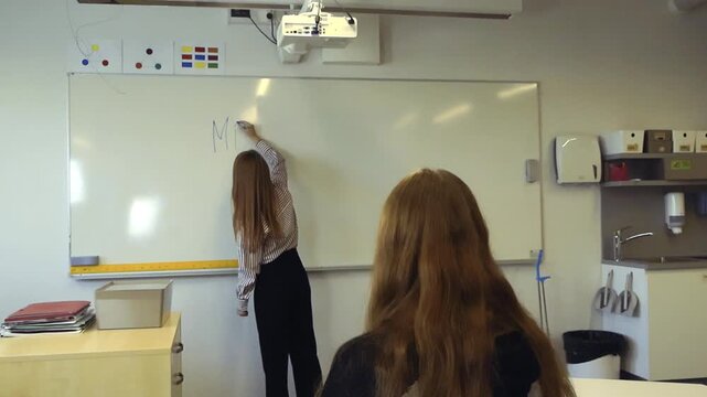 Young female teacher writing about the Middle Eastern conflict on a whiteboard during a history or political science lesson, with a student watching attentively from her desk