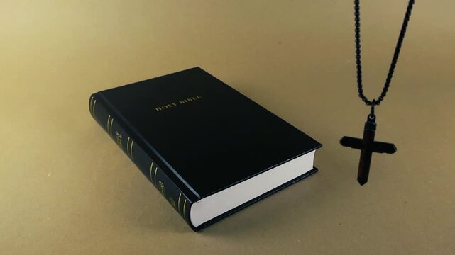 Black Holy Bible resting on a light brown surface with a black christian cross necklace swinging next to it. Religious concept representing faith, spirituality, and belief in God