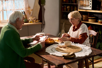 Senior Caucasian couple preparing food and reading newspaper at kitchen table. Image suitable for...