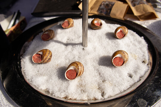 Metal tray filled with coarse salt, holding snail shells stuffed with seasoned filling. Culinary presentation of escargot at a street food festival
