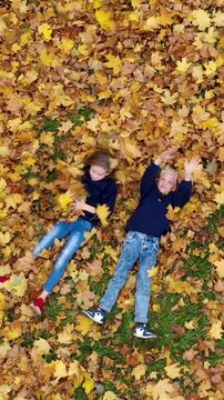 Vertical video: an aerial view of a happy boy and girl lying on the ground and tossing yellow autumn leaves into the air in a park. Drone footage: two children having fun. 9:16 format for social media