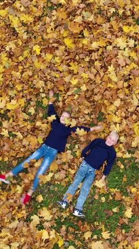 Vertical video: Aerial view of a happy boy and girl lying on ground and making leaf angels in autumn park. Drone shot of two kids moving arms and legs in yellow foliage. 9:16 format.