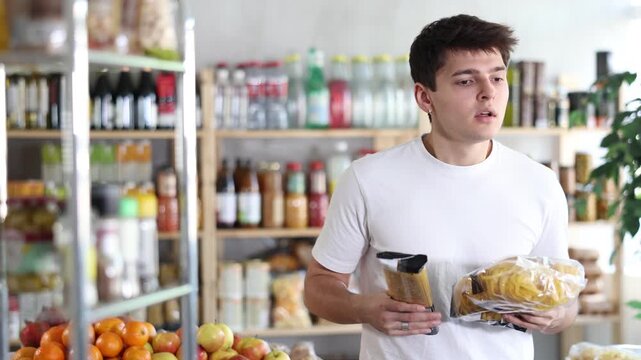 Guy choose and buy pasta in store. Market visitor consider and make choice, buy pasta garnish products. Client hesitates before buying, choose noodles, reads labels on package 