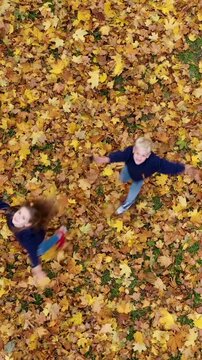 Vertical video: Aerial view of a happy boy and girl spinning around and throwing yellow autumn leaves up in park. Drone shot of two kids having fun. 9:16 social media format.