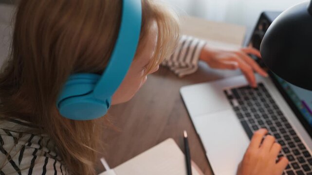 Teenage girl wearing headphones using laptop at home for online learning, studying at desk and engaging with digital educational content, representing modern education, technology and remote learning