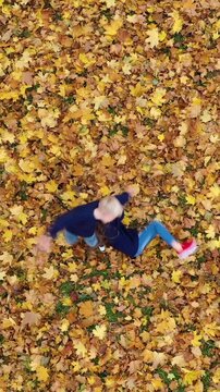 Vertical video: Aerial view of a happy boy and girl spinning around and throwing yellow autumn leaves up in park. Drone shot of two kids having fun. 9:16 social media format.