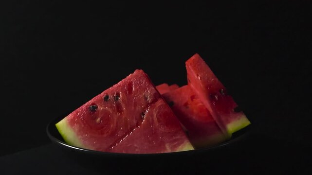 Juicy, ripe watermelon slices with seeds spinning on a black plate against a dark background. Fresh, sweet fruit presentation perfect for healthy eating, summer concepts, and vitamin rich diets