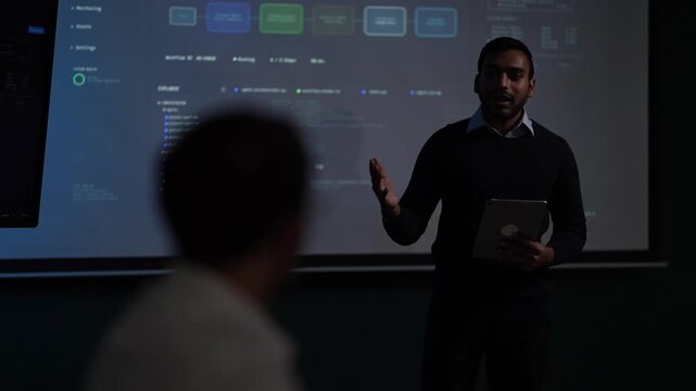 A technical founder addressing his team gestures with an open hand mid sentence while a listener sits silhouetted in the foreground inside a dark AI startup office.